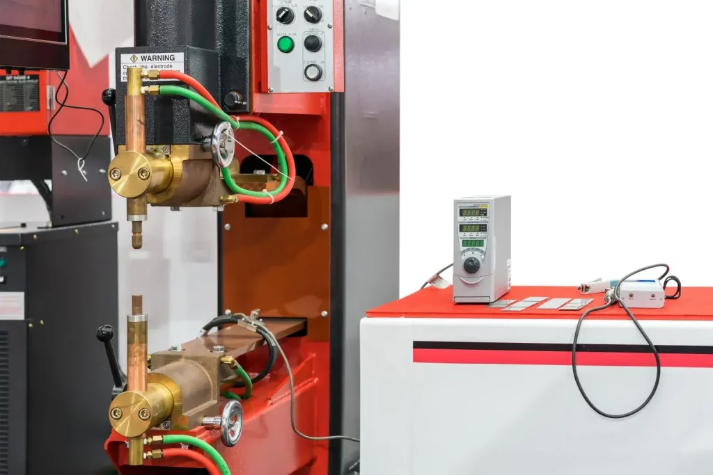 Nahaufnahme einer Industriemaschine mit Messingventilen, grünen und roten Schläuchen, Bedienknöpfen und einem digitalen Gerät auf einem rot-weißen Bedienfeld vor einem schlichten weißen Hintergrund.