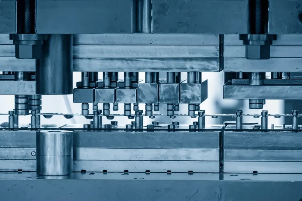 Nahaufnahme einer industriellen Metallstanzmaschine mit verschiedenen Bolzen, Platten und Bauteilen, die feinmechanische und fertigungstechnische Geräte in einer Fabrikumgebung zeigt.